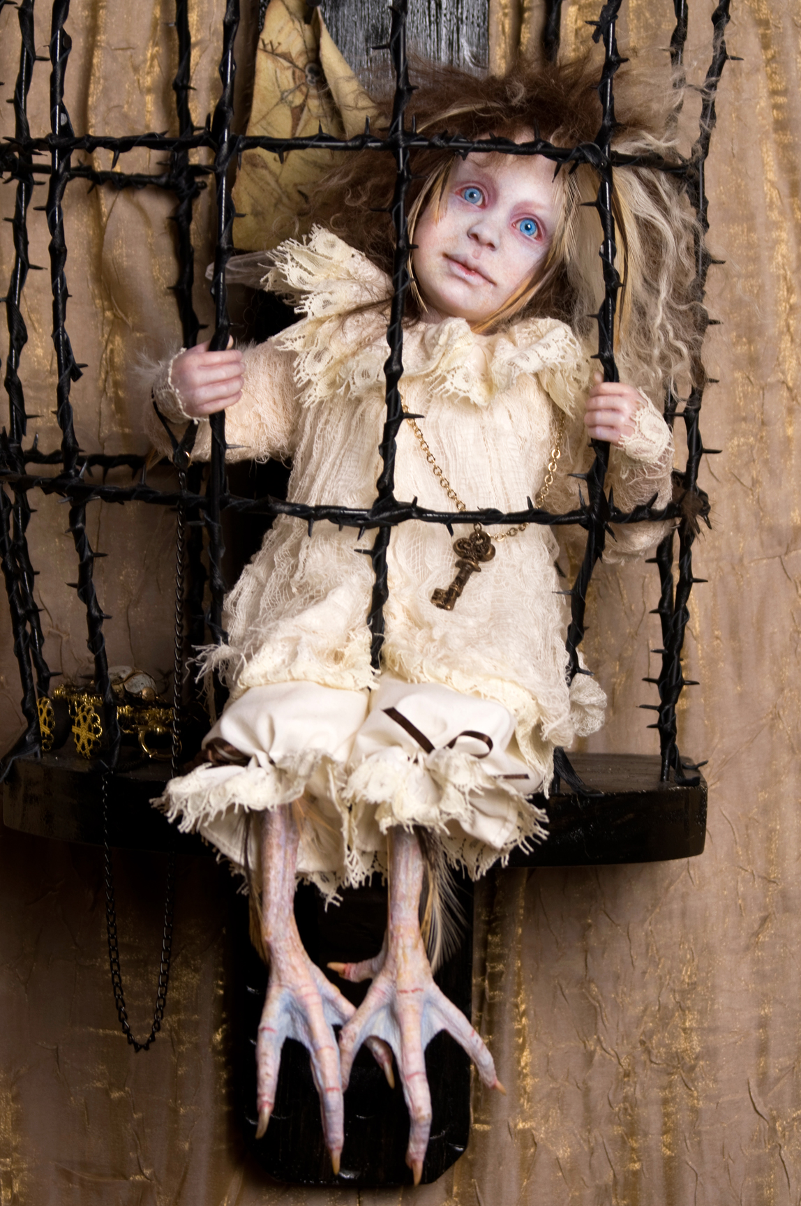pale blue eyed blond feathered gothic artdoll in a white lace nightie with taxidermied bird feet sits in a black thorn cage with a large key on a chain around his neck.