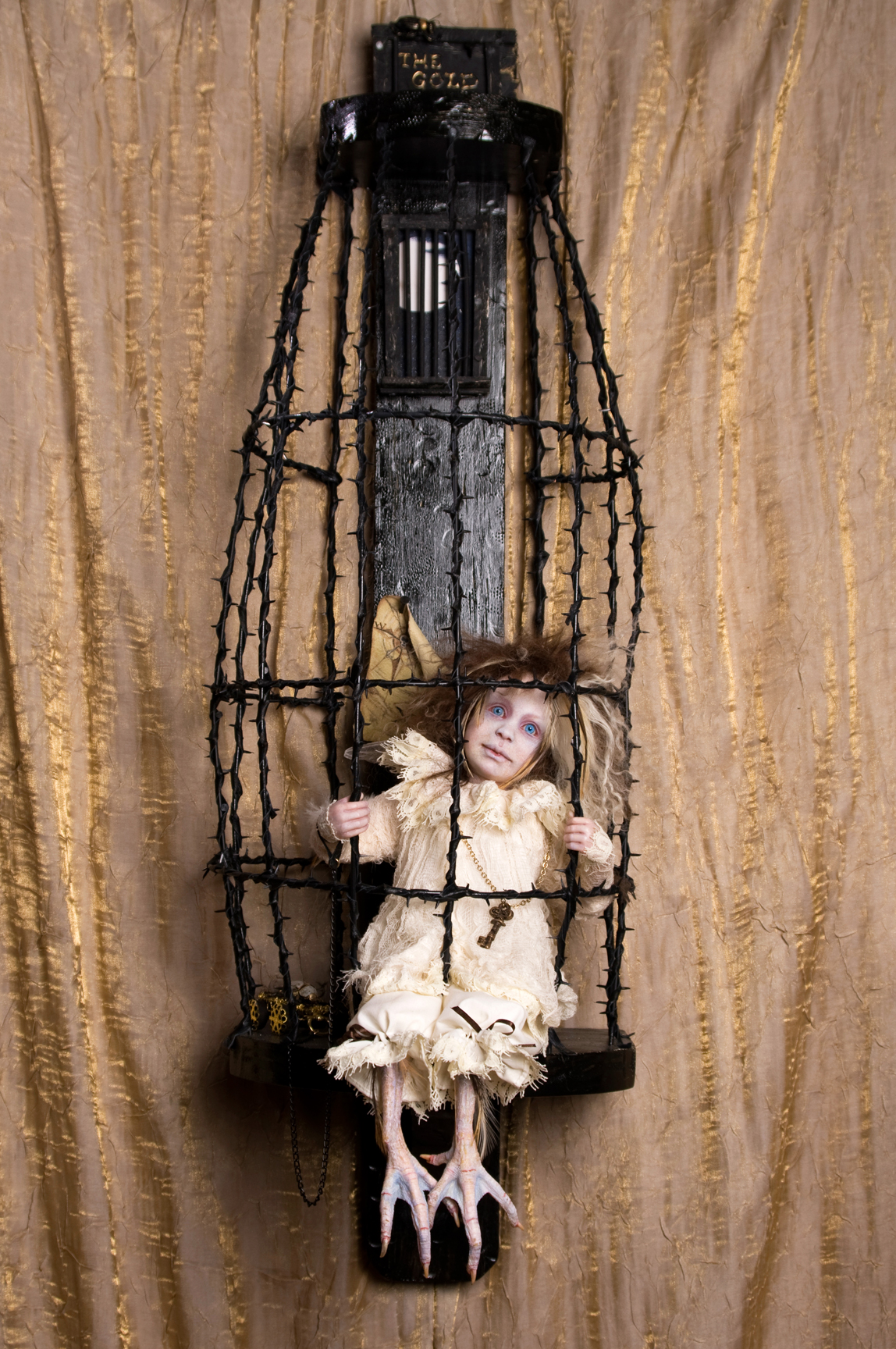 pale blue eyed blond feathered gothic artdoll in a white lace nightie with taxidermied bird feet sits in a black thorn cage with a large key on a chain around his neck.