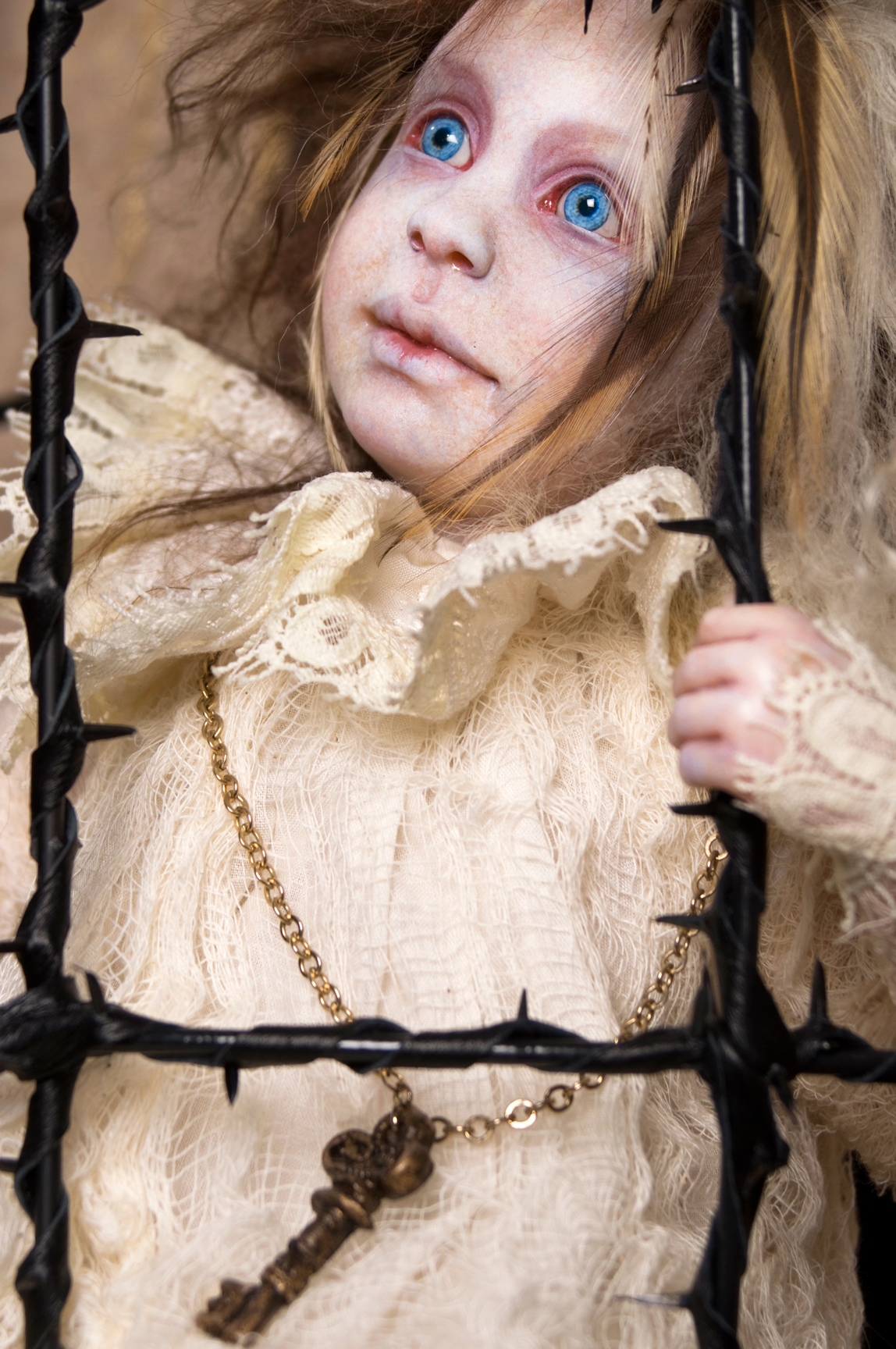 pale blue eyed blond feathered gothic artdoll in a white lace nightie sits in a black thorn cage with a large key on a chain around his neck.