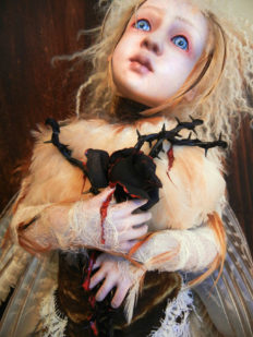close-up of blue eyed blond feathered artdoll clutching a black rosebush branch with thorns and a black rose dripping blood
