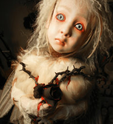 close-up of blue eyed blond feathered artdoll clutching a black rosebush branch with thorns and a black rose dripping blood