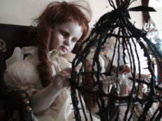 gothic artdoll pale with red ringlets wearing a white dress holding a black barbed birdcage in her lap peering in at a tiny sculpted bird.