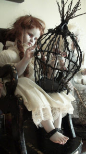 gothic artdoll pale with red ringlets wearing a white dress with bound feet in spiked cuffs holding a black barbed birdcage in her lap peering in at a tiny sculpted bird.