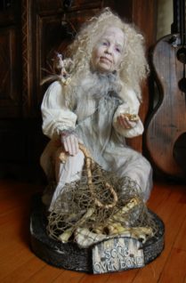 old woman artdoll representing the Crone she is seated in a white nightgown and holds a netted bag of animal bones in one hand and a baby bird skeleton in the other hand
