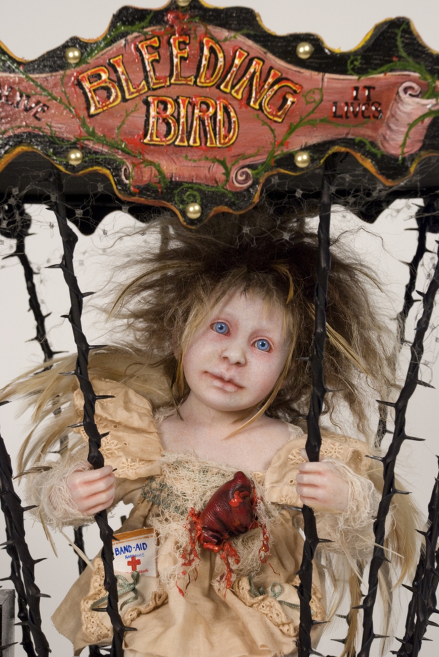 close-up of artdoll with exposed bleeding heart sitting in a throned cage on a circus cart handpainted sign reads Bleeding Bird