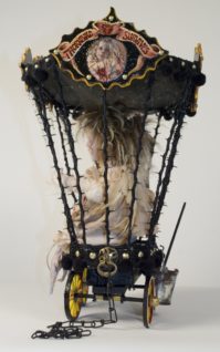 Side view of taxidermy artdoll with bird feet sitting in a caged circus cart sideshow hand-painted freakshow signs