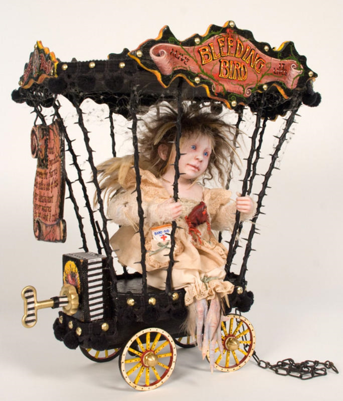 a taxidermy assemblage art doll with bird feet sits in a circus cage on a cart music box with hand painted signs sideshow freak show bleeding heart