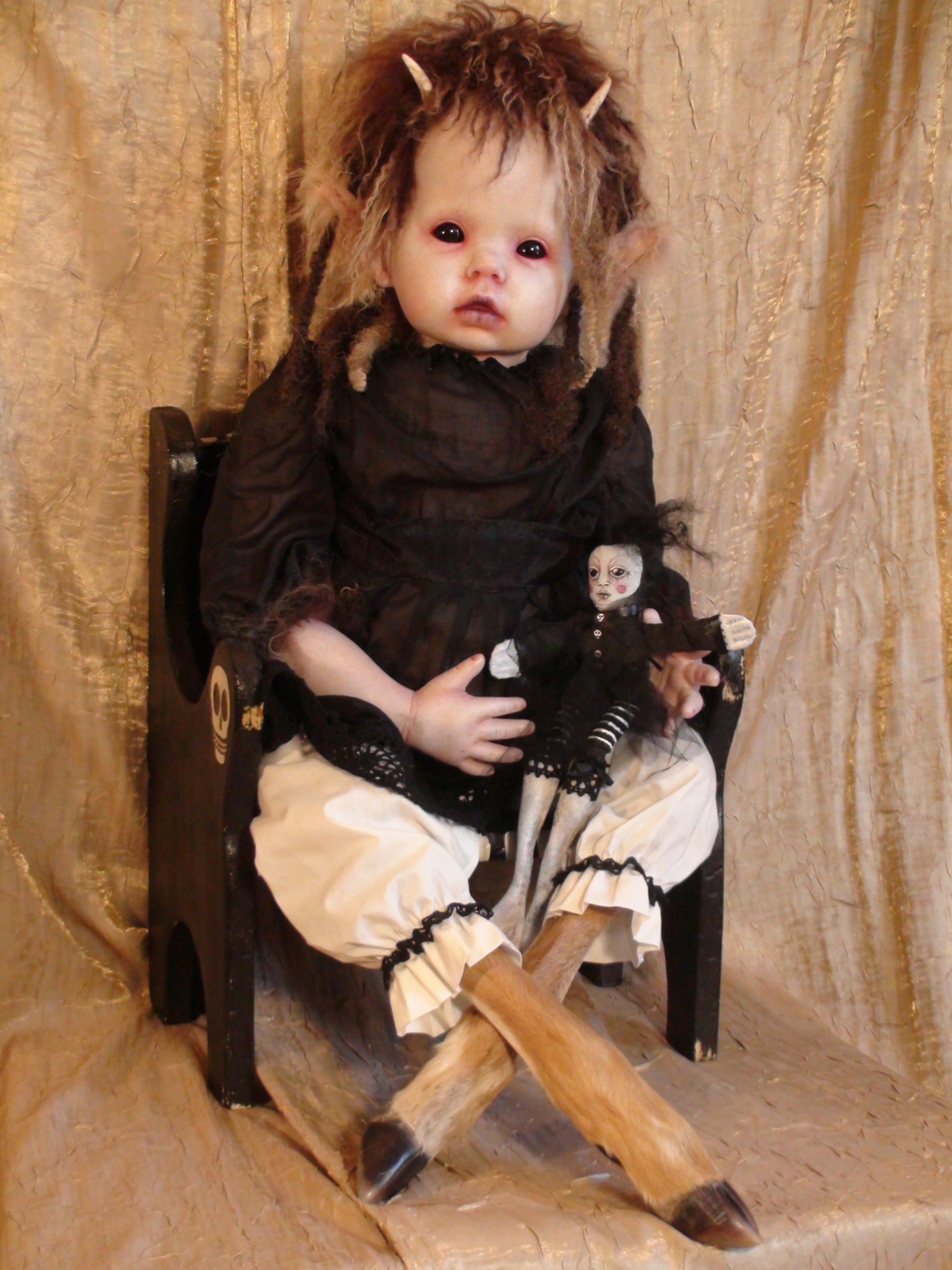 seated gothic artdoll with taxidermy deer legs she is pale with short brown hair, elf ears, horns she wears a black top and white pants and holds a little gothic cloth doll in her lap