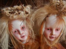 close-up two-headed twin artdolls heads together pale with blond feathered hair bird nests on heads