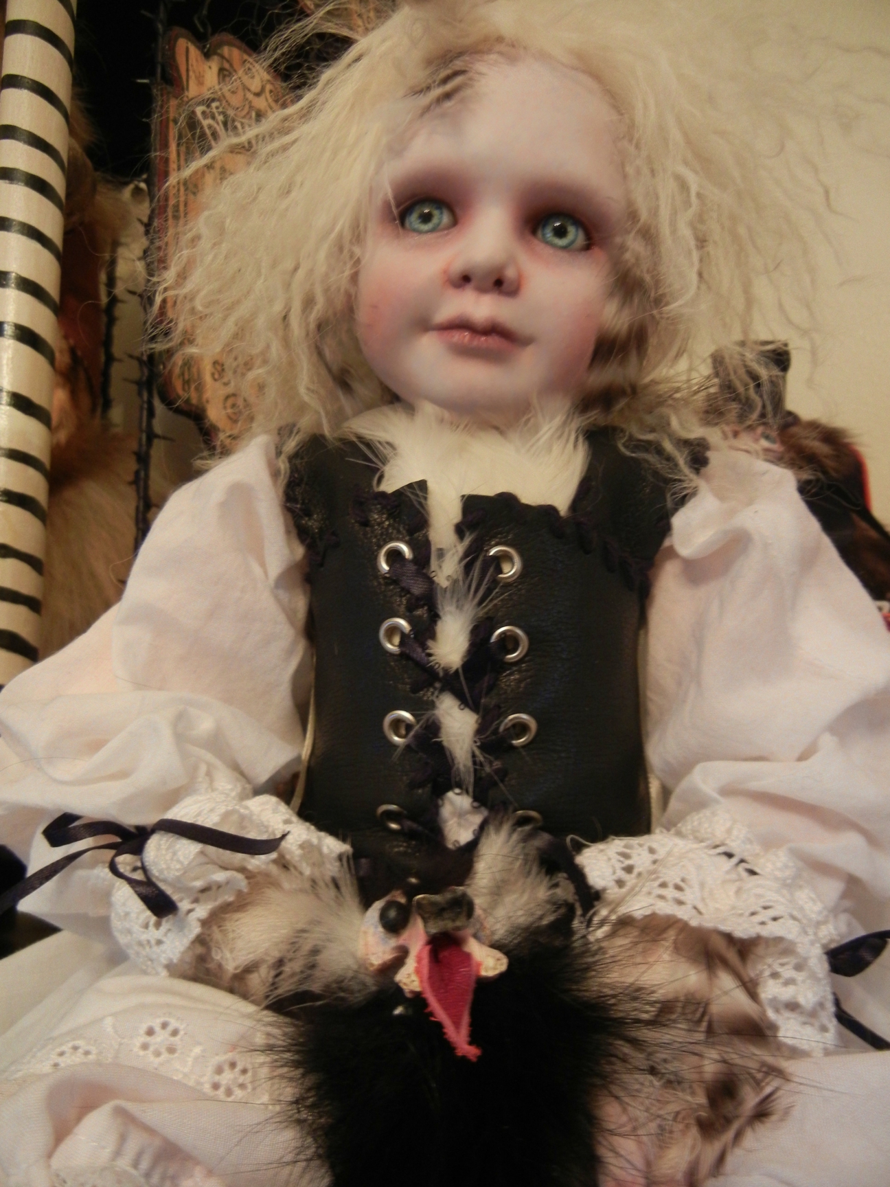 seated gothic artdoll pale with light eyes and blond hair wearing black beret, white shirt with lace cuffs, leather vest, taxidermy chicken feet holds a feathered pet creature in his lap