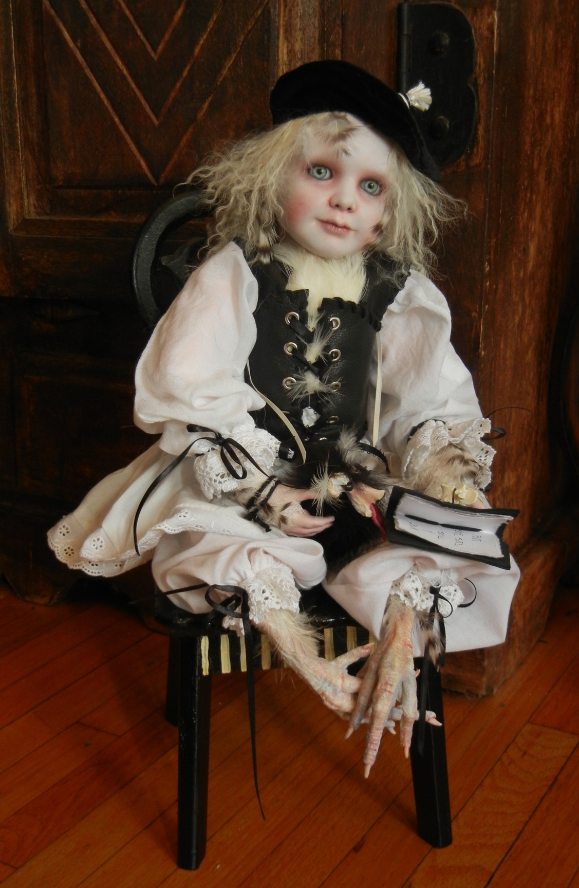 seated gothic artdoll pale with light eyes and blond hair wearing black beret, white shirt with lace cuffs, black leather vest, taxidermy chicken feet holds a feathered pet creature in his lap