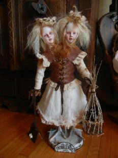 gothic two-headed twin artdolls with blond hair and taxidermied bird feet dressed as peasants with leather vest and white skirt nests on their heads holding bird cage made of sticks