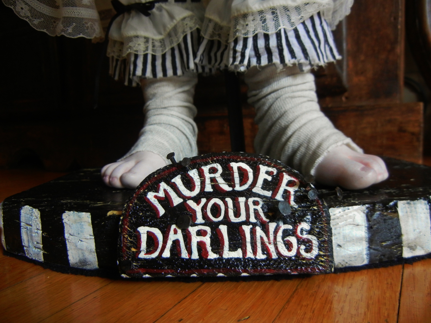 dollfeet wrapped in white material with black and white stripe pant cuffs standing on hand painted wooden platform with sign that reads Murder Your Darlings