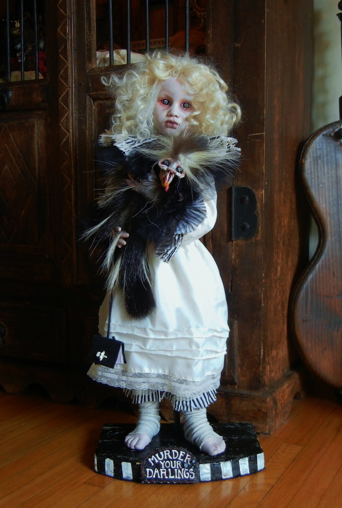 pale gothic artdoll with blond ringlets, skulls in her black eyes, wearing a white dress, carrying black feathered creature sculpture and standing on a painted wooden platform