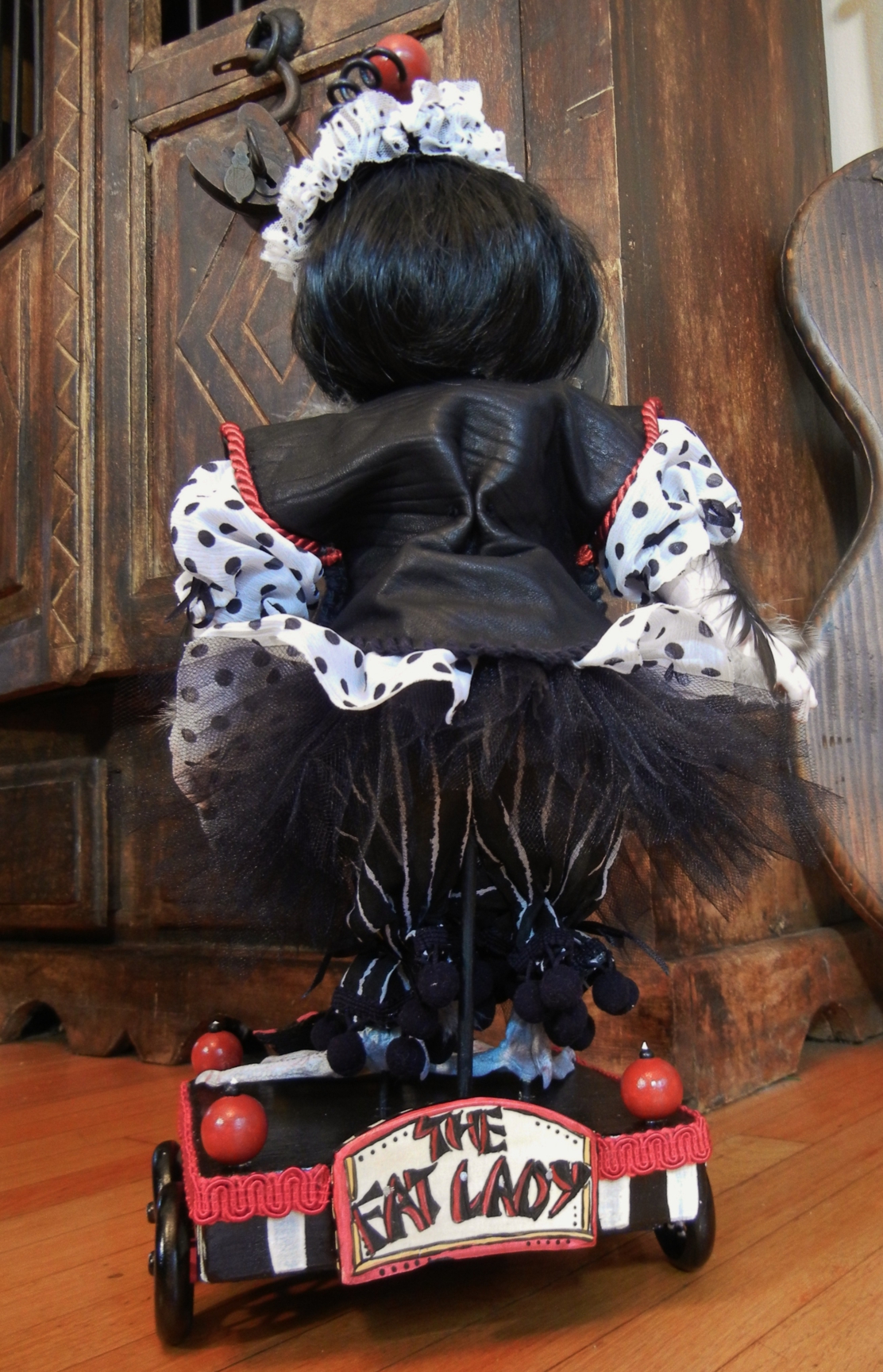 Asian Chinese girl doll with black bob wearing black and white polka-dot dress has feathers and taxidermy bird webbed duck feet standing on hand-painted wooden circus cart