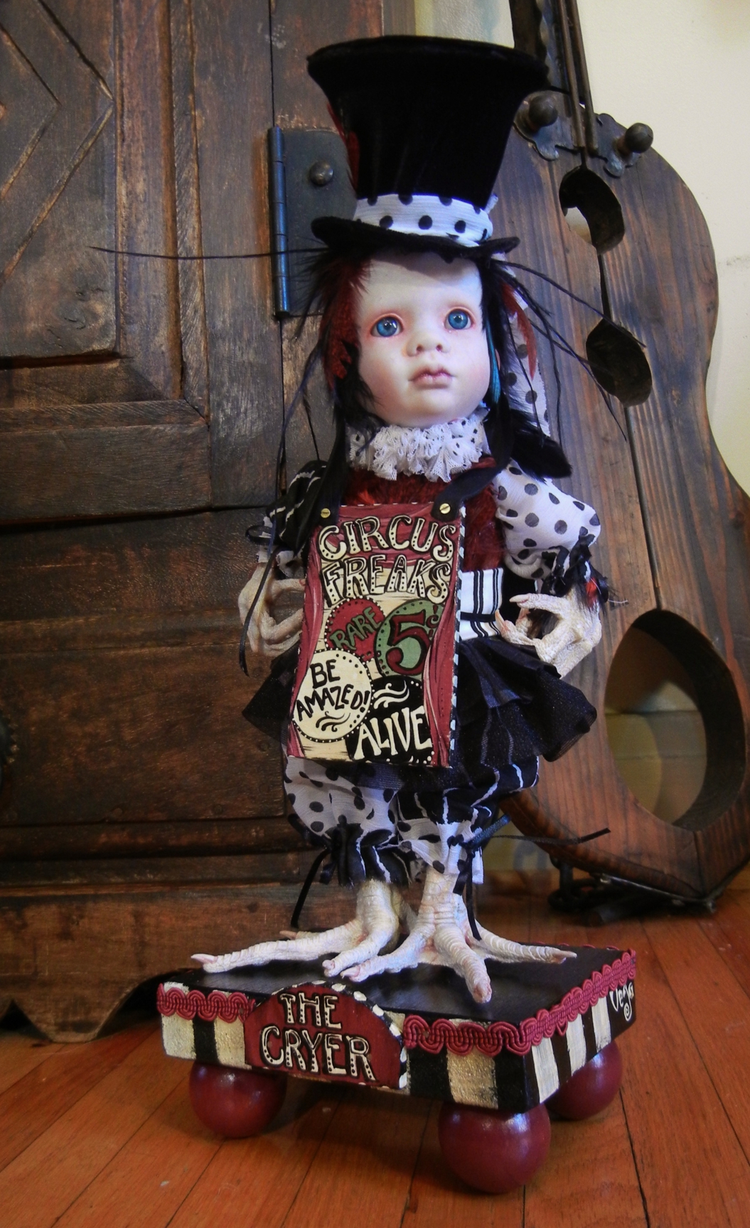 gothic feathered mixed media artdoll with taxidermy birdfeet. Sideshow-themed circus cryer wearing hand-painted wooden signboard, black velvet tophat, black and white doll clothes, stands on a hand-painted wooden platform
