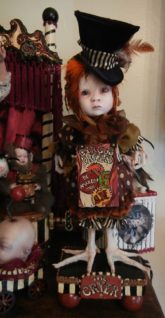 gothic feathered mixed media artdoll with taxidermy birdfeet. Sideshow-themed circus cryer wearing hand-painted wooden signboard, black velvet tophat, polka dot doll clothes, stands on a hand-painted wooden platform