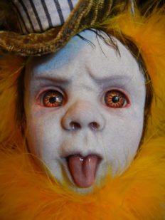 close-up porcelain babydoll repaint head with tongue sticking out, orange eyes, brown top hat, yellow feathers around his face