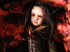 miniature goth doll pale with black hair and black clothes in front of red background black branches