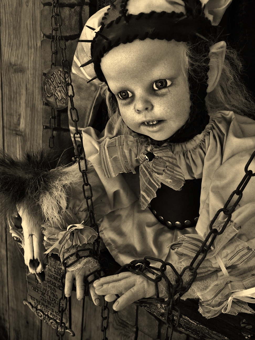 black and white image close-up gothic artdoll wearing white jester cap stands in window with chains