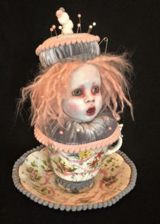 pale porcelain doll head gothic repaint wild strawberry blond hair and a velvet pincushion hat sits on a gray velvet cushion with pins in it in a vintage floral teacup