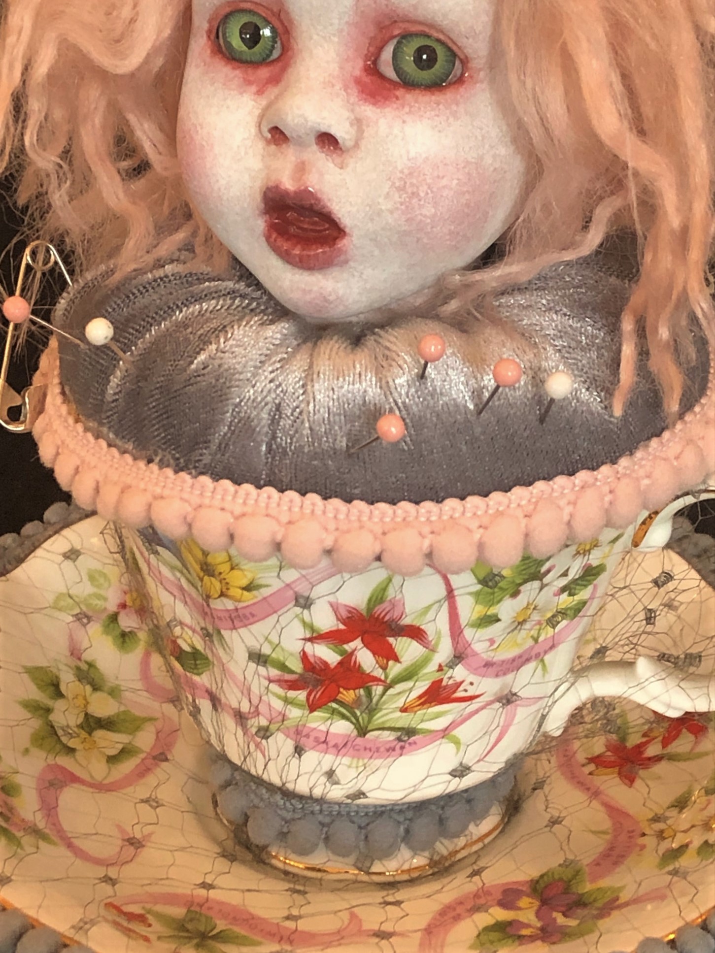 close-up of pale porcelain doll head gothic repaint wild strawberry blond hair sits on a gray velvet cushion with pins in it in a vintage floral teacup