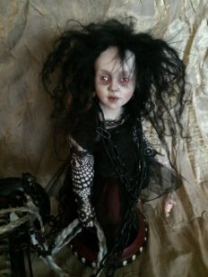miniature pale goth doll with black hair and lace black sleeves wearing read and black