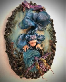 resin encased mixed media assemblage of a doll faced blue fish rising out of the water to see a bejeweled beetle.