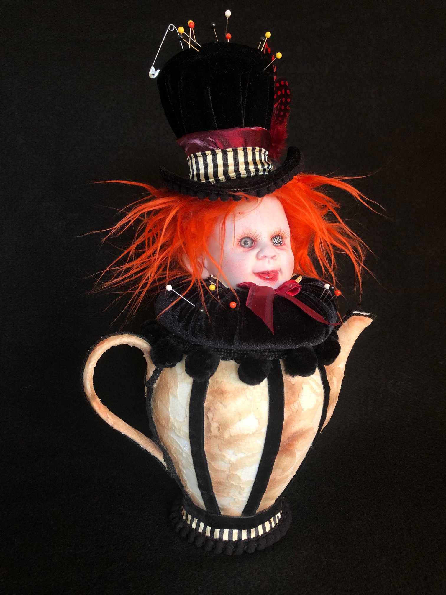 mad hatter themed pincushion porcelain doll head with bright orange hair and velvet tophat sits on a black velvet cushion set in a lace-covered porcelain teapot