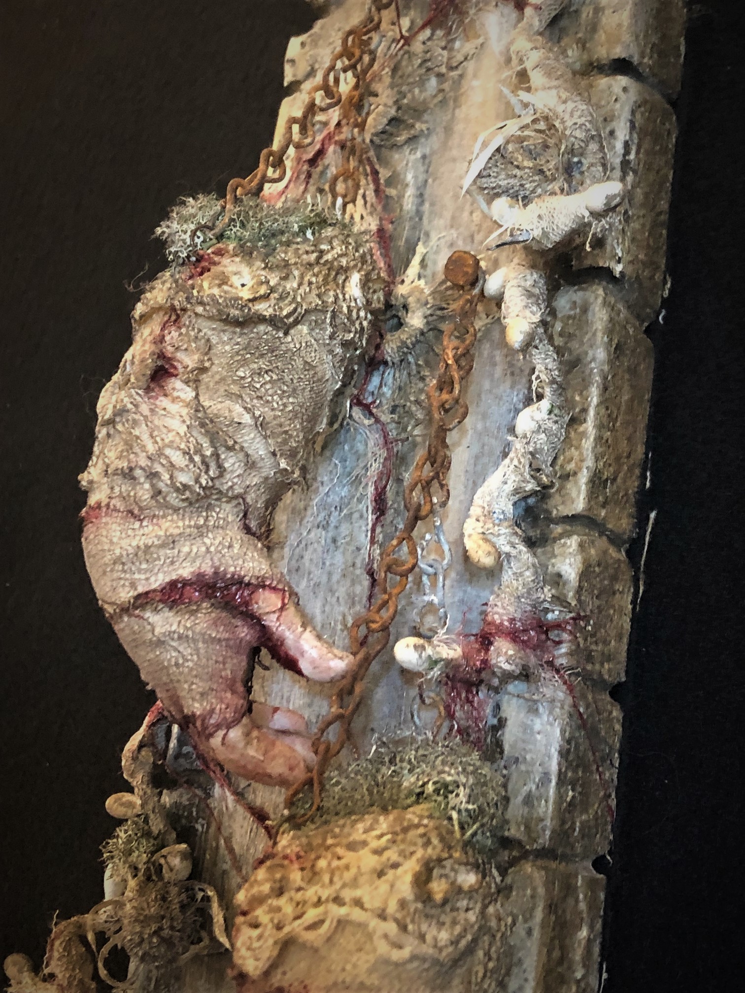 close-up of mixed media assemblage on board with severed, cracked and bleeding hands held up by chain and hanging from nails