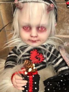 creepy baby holiday elf doll with red eyes white hair and fur dressed in stripes with horns, canine paws, and a reptilian tail.