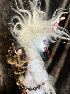 close up leering white albino krampus art doll with his tonue out white hair and fur, horned, with a basket with a baby on his back