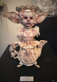 white mixed media assemblage with a cat eared babydoll head and a porcelain teapot with a skeletal handle
