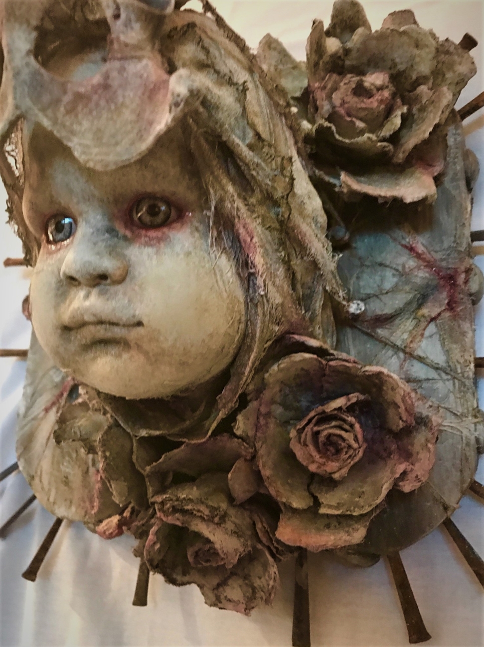 close-up of mixed media assemblage plaque repainted babydoll head with a bone headdress and flowers and nails mounted on wood
