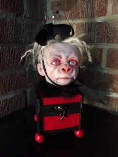 pale, albino gorilla face doll head with red open eyes and blond hair wearing a black pin cushion with a red tassel on his head set on top of a burgundy and black wooden box