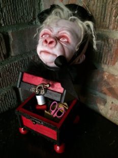 pale, albino gorilla face doll head with red open eyes and blond hair wearing a black pin cushion with a red tassel on his head set on top of an open burgundy and black wooden box with sewing notions inside the box