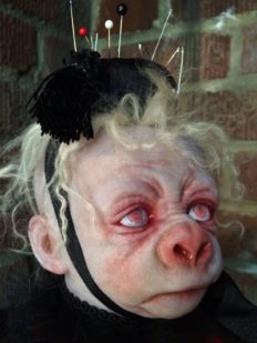 close-up pale, albino gorilla face doll head with red open eyes and blond hair wearing a black pin cushion with a red tassel on his head