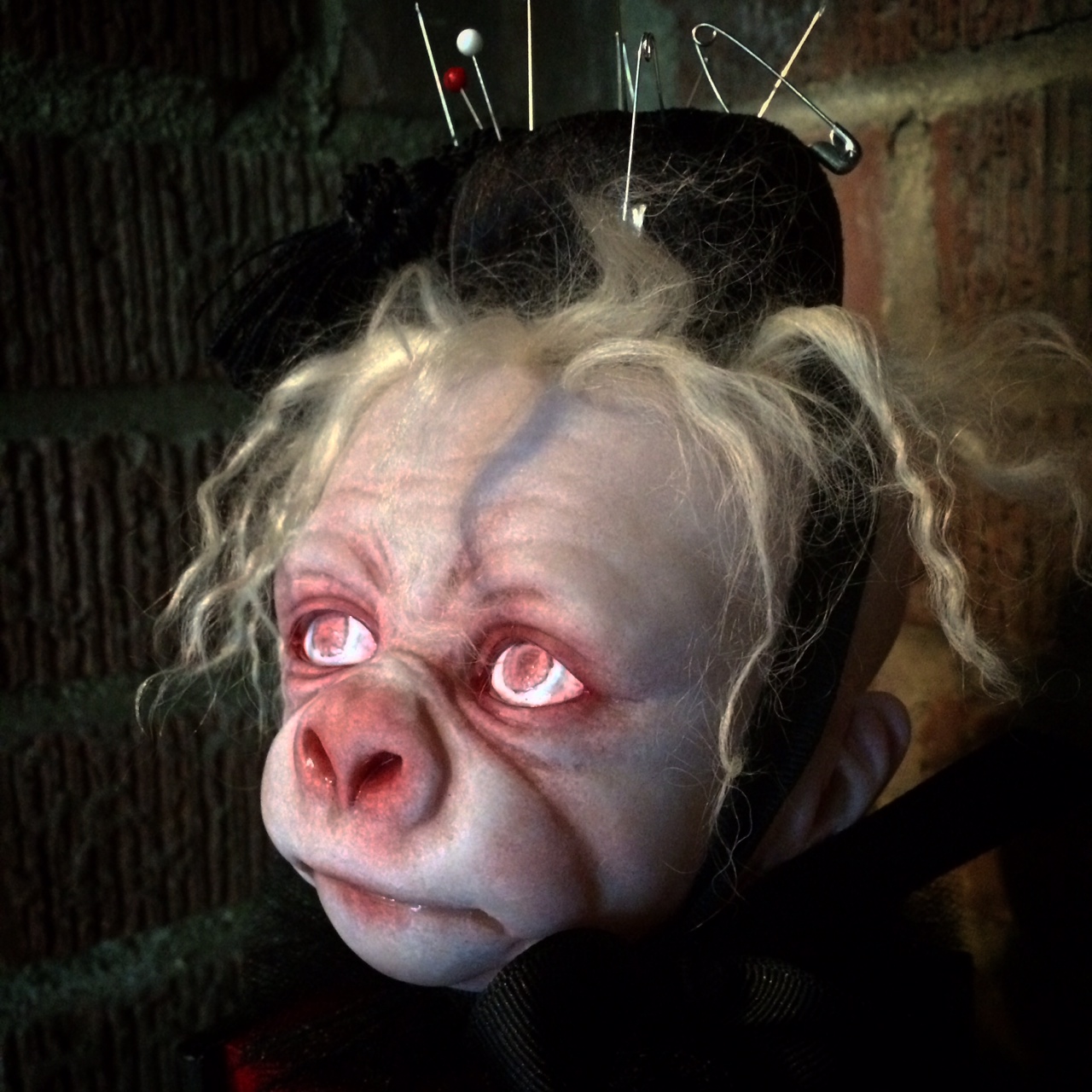 close-up pale, albino gorilla face doll head with red open eyes and blond hair wearing a black pin cushion with a red tassel on his head