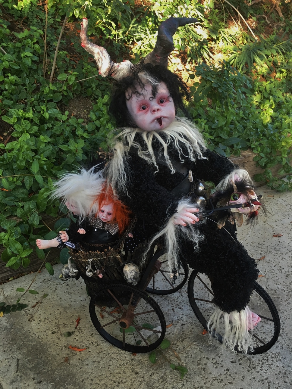 baby krampus artdoll with fur and horns is riding on a vintage tricycle with child dolls in his basket