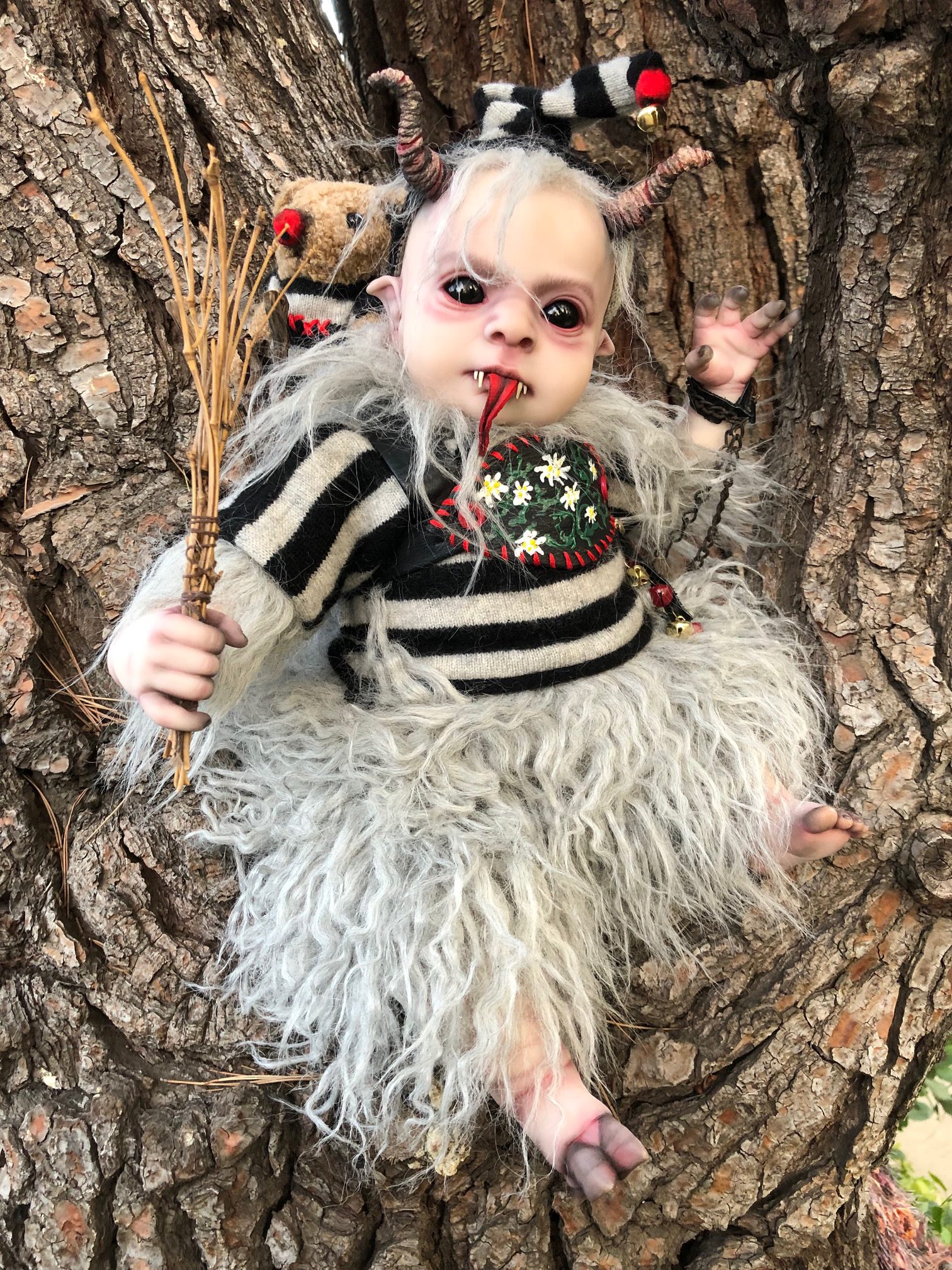 mixed media artdoll creepy evil babydoll elf with horns, black eyeballs, pointed elf ears, tongue sticks out, faux fur collar, striped cap and sweater, cloven hoof foot, curly tail, faux fur legs and teddy bear on his back