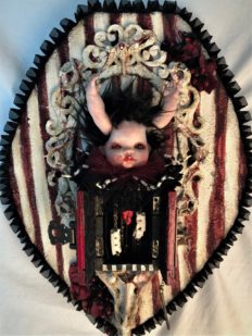 gothic red black and white mixed media assemblage artpiece plaque with a repaint porcelain babydoll head with arms for horns, striped open cabinet, surrounded by vertebrae on textile covered wooden board
