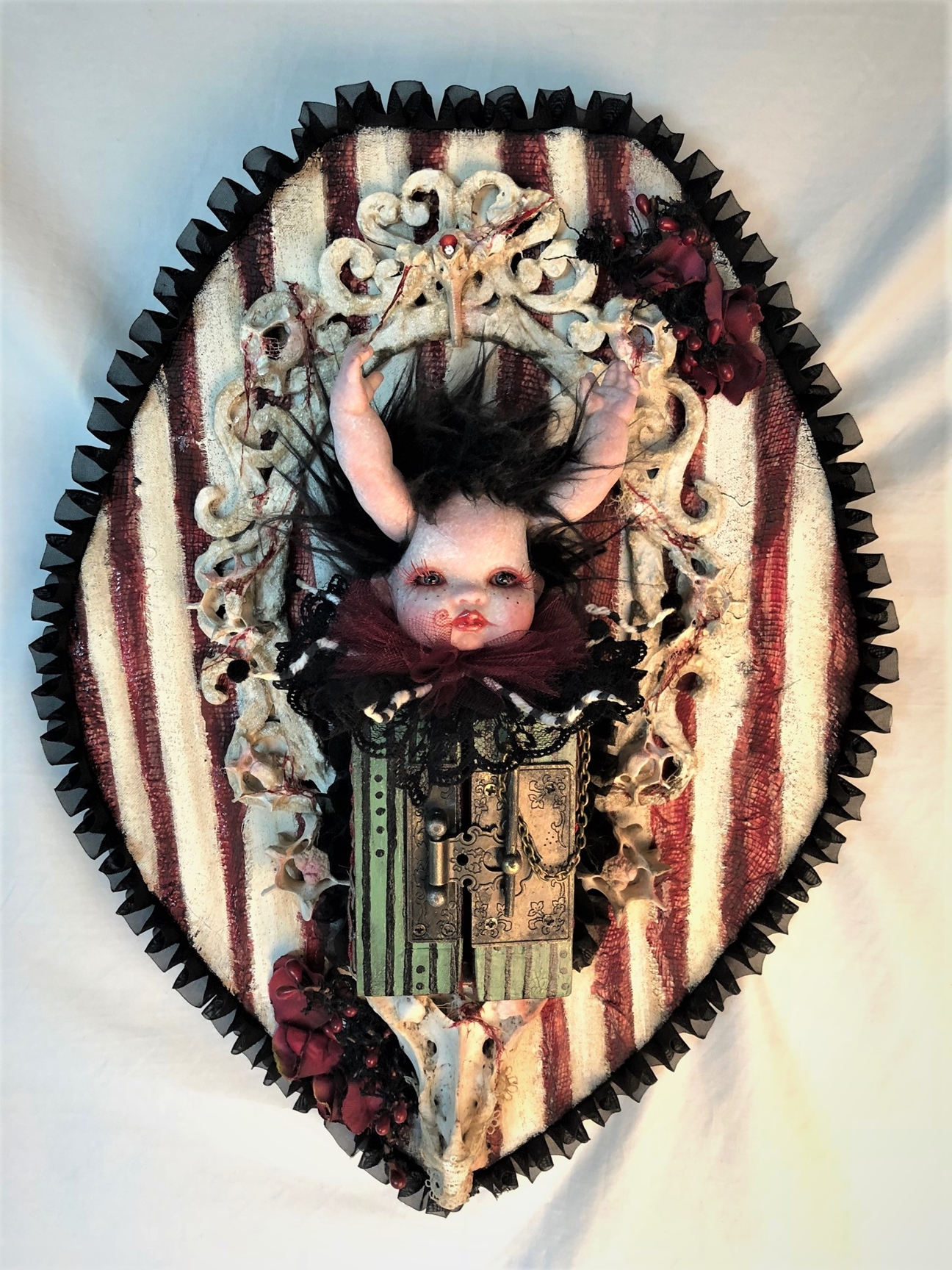 gothic red black and white mixed media assemblage artpiece plaque with a repaint porcelain babydoll head with arms for horns, striped closed cabinet, surrounded by vertebrae on textile covered wooden board