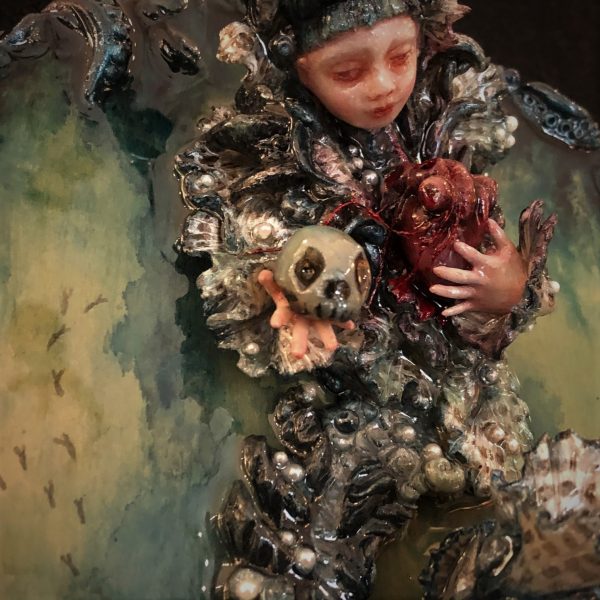 close-up of resin encased mixed media assemblage of a doll-faced lion fish creature holding a skull and a heart.