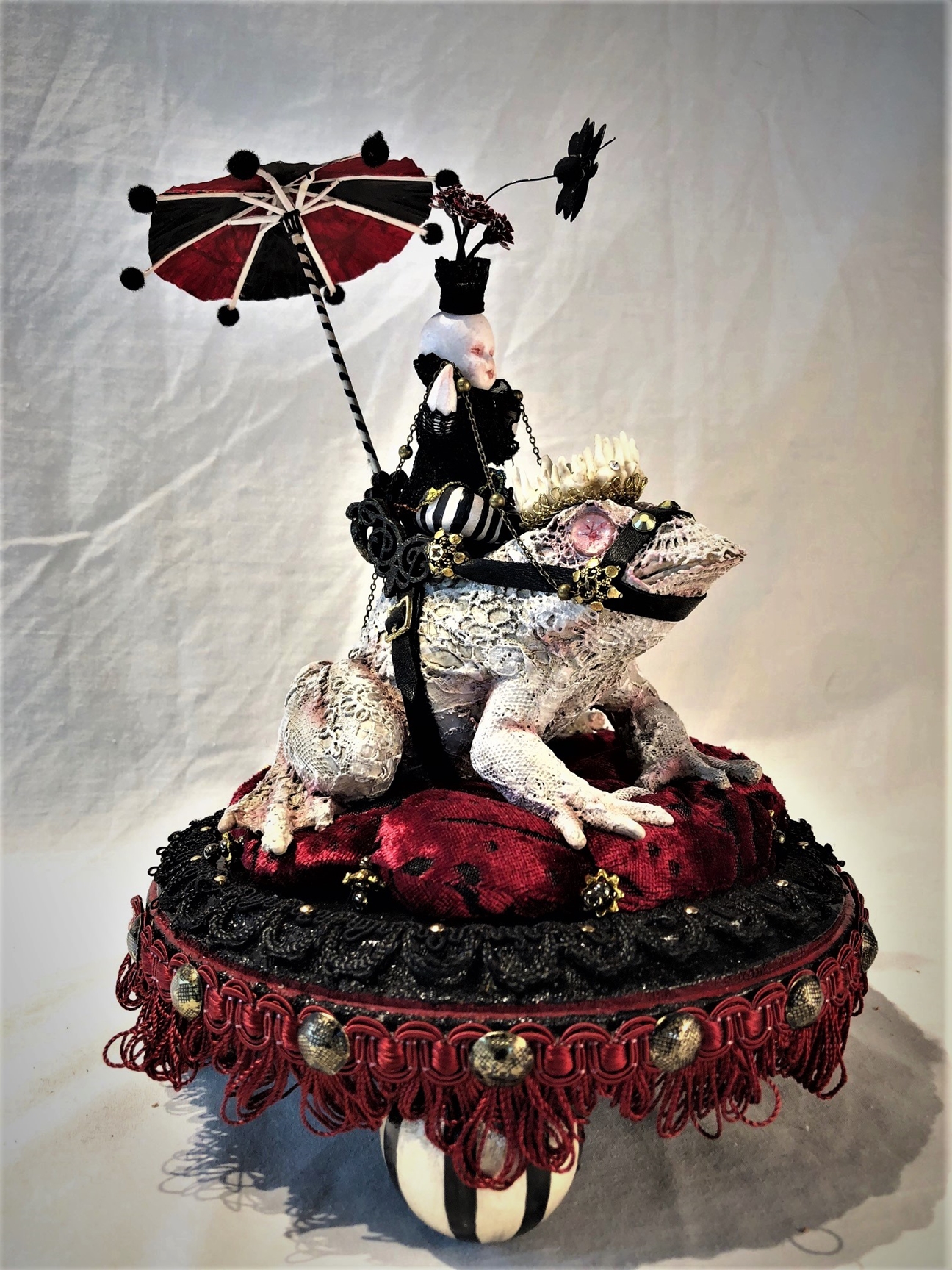 mixed media striped miniature gothic circus performer under a parasol rides a lace-covered vintage toy frog with a crown made of teeth on fabric and fringe decorated wooden platform