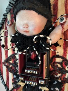 close-up red, white and black striped mixed media gothic circus-themed artpiece of a doll holding her own bloody eyeballs with a small cabinet holding a heart for her torso on striped textile covered wooden board