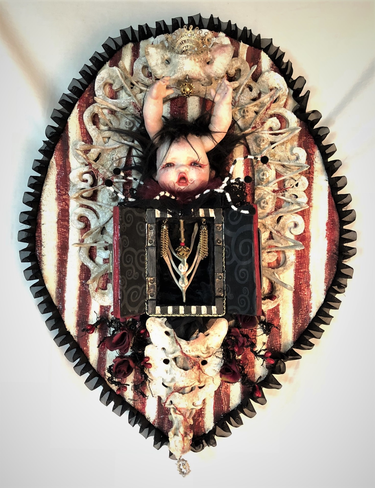 gothic red black and white mixed media assemblage artpiece plaque with a repaint porcelain babydoll head with arms for horns, striped open cabinet, vertebrae, surrounded by wishbones on a painted wooden board