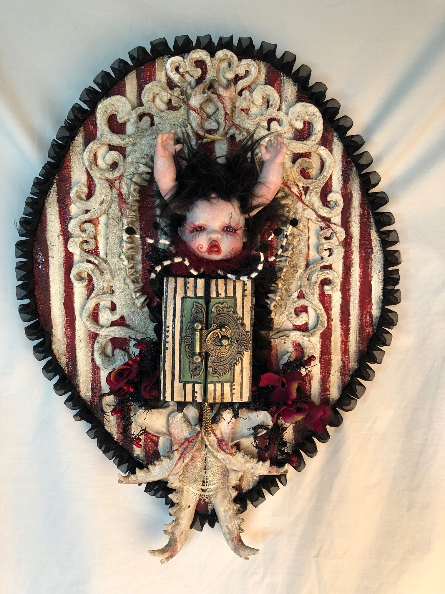 mixed media assemblage wall plaque with gothic repaint porcelain doll head arms for horns, closed striped cabinet, jawbones on striped textile covered wooden board