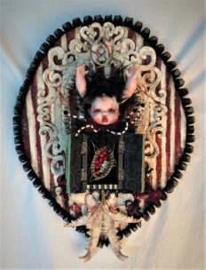 mixed media assemblage wall plaque with gothic repaint porcelain doll head arms for horns, open cabinet revealing tiny basket, jawbones on striped textile covered wooden board