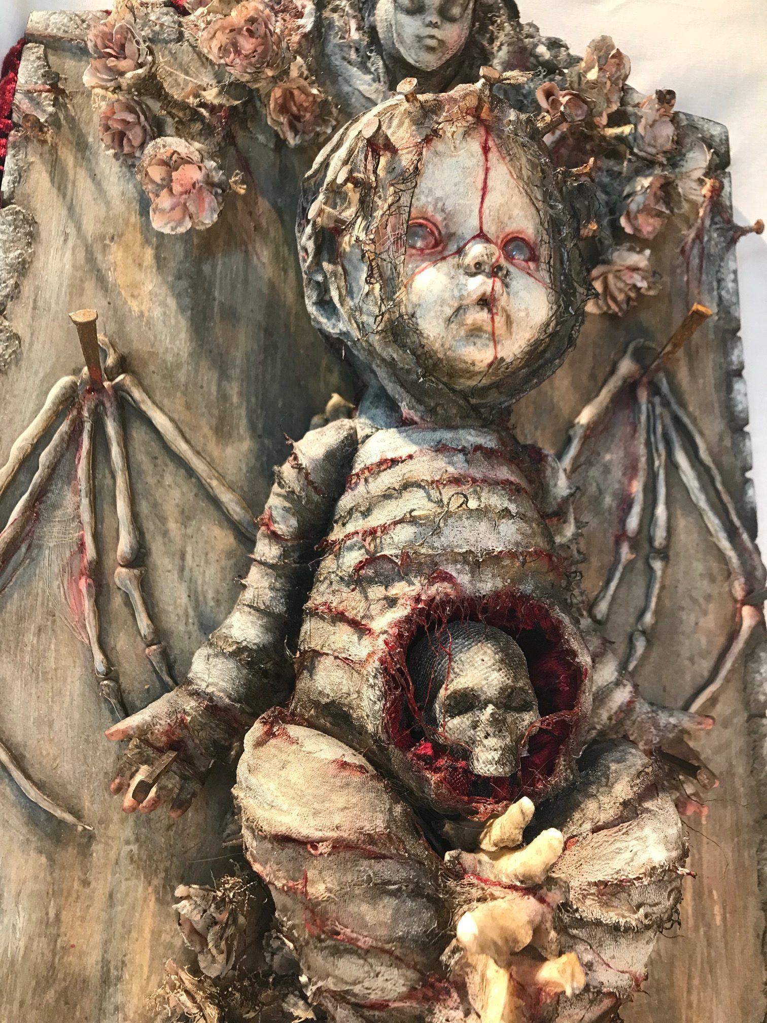 close-up of mixed media assemblage plaque bloody mummified babydoll with skeleton wings and a skull in the stomach