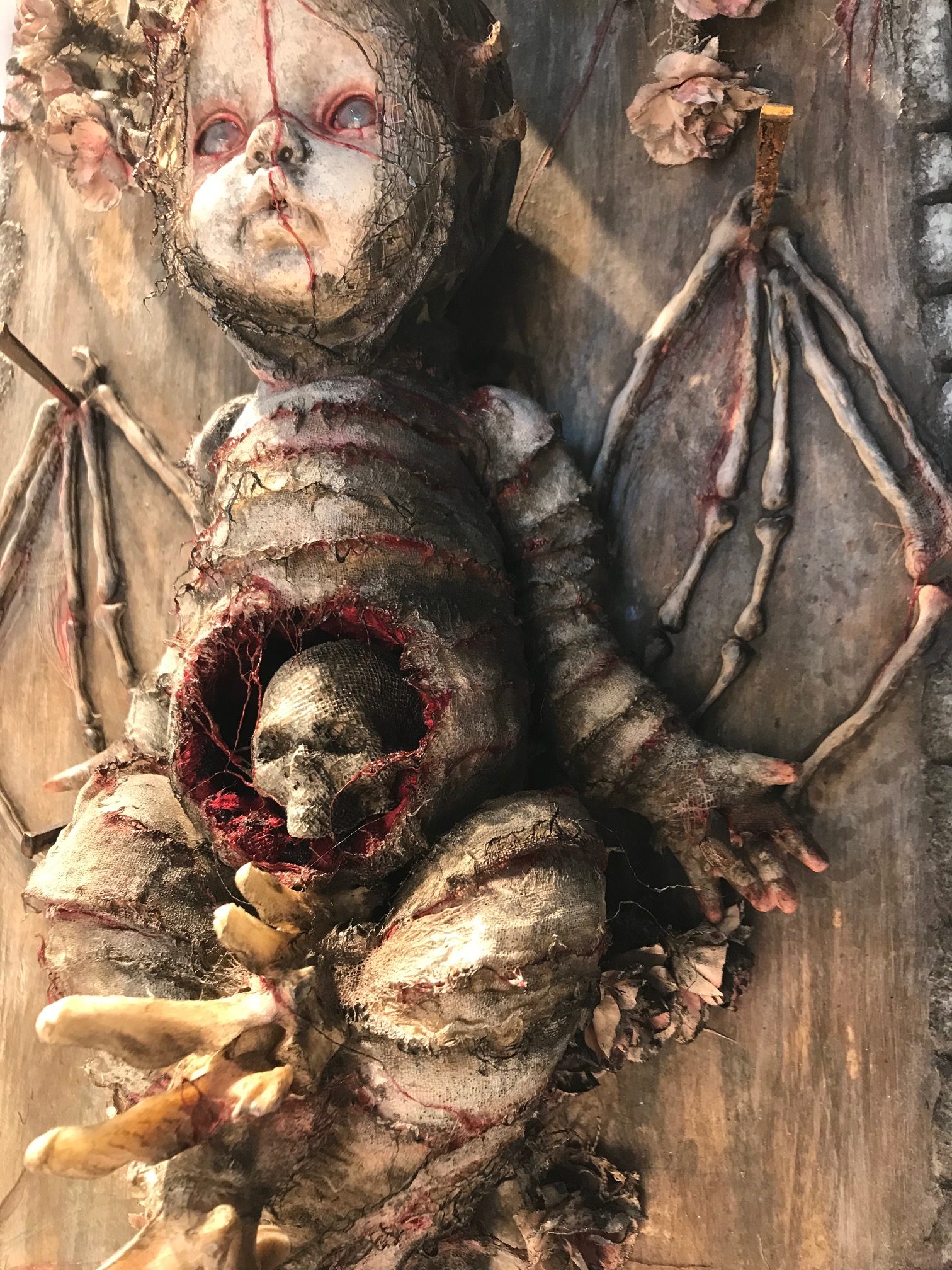 close-up of mixed media assemblage plaque bloody mummified babydoll with skeleton wings and a skull in the stomach